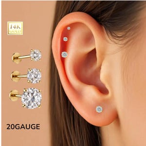 May include: Gold-tone stud earrings with clear, sparkling cubic zirconia stones. The earrings are displayed next to an ear with multiple piercings. The text "20 GAUGE" is visible. The earrings are labeled "14K GOLD".