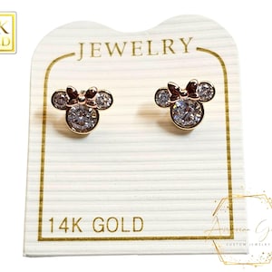 Puede incluir: Un par de pendientes de oro de 14 quilates con un diseño de Mickey Mouse. Los pendientes están engastados con piedras de circonita cúbica transparentes y tienen un acabado en oro rosa. Los pendientes se muestran en una tarjeta blanca con las palabras "Joyería" y "Oro de 14 quilates" impresas en ella.