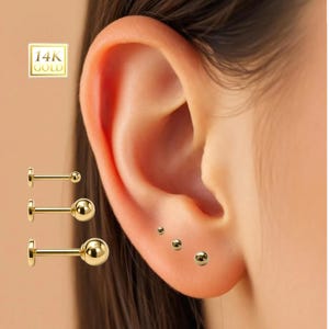 May include: Close-up of an ear adorned with multiple gold ball stud earrings. Three larger earrings are displayed on the left, with the text "14K GOLD" in the upper left corner. The ear has three smaller earrings.