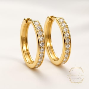 Puede incluir: Un par de pendientes de aro dorados con una fila de pequeñas piedras preciosas transparentes a lo largo del borde exterior. Los pendientes se muestran sobre una superficie blanca, con luz suave. Un diseño de joyería clásico.
