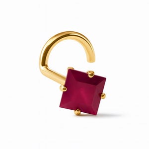 May include: A gold-colored nose ring featuring a square, deep red gemstone. The gemstone is held in place by four small gold prongs. The nose ring has a curved, hook-like design.