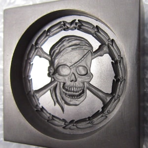 Könnte beinhalten: Eine schwarze und graue Form mit einem erhabenen Design eines Schädels und Knochen mit Augenklappe und Bandana. Der Schädel und die Knochen sind von einem Kranz aus Blättern umgeben.