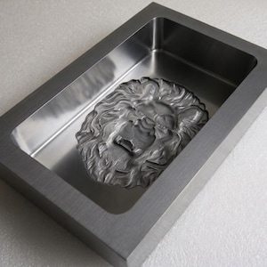 Puede incluir: Una bandeja rectangular de metal con un acabado cepillado en el borde exterior y una base interior pulida y reflectante. Centrado en el fondo de la bandeja hay un relieve tridimensional esculpido de una cabeza de león con melena detallada.