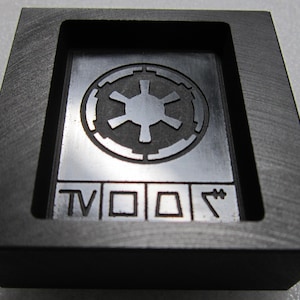 Puede incluir: Un marco rectangular negro que contiene una placa de plata metálica. La placa presenta un símbolo circular del emblema imperial en la parte superior y cuatro caracteres Aurebesh distintos grabados en una fila horizontal en la parte inferior.