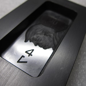 Może przedstawiać: Prostokątny, ciemnoszary metalowy przedmiot z zagłębionym obszarem zawierającym szczegółowy profil twarzy. Na powierzchni wytrawiono cyfrę "4". Obiekt umieszczony jest w większej, pasującej ramie.