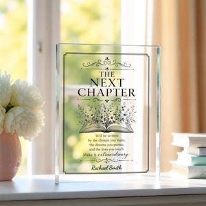 May include: A rectangular clear acrylic plaque featuring black printed text that reads The Next Chapter, followed by a floral illustration of an open book and the quote Will be written by the choices you make, the dreams you pursue, and the lives you touch. Make it extraordinary. Rachael Smith.