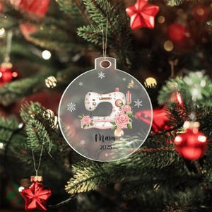May include: Clear, round Christmas ornament featuring a sewing machine design with pink roses and snowflakes. The ornament includes the name "Mary" and the year "2025". The ornament hangs from a gold string against a blurred Christmas tree background.