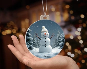 Puede incluir: Un adorno navideño redondo con un muñeco de nieve en una escena de bosque nevado. El muñeco de nieve sonríe y lleva un sombrero negro. El adorno está personalizado con el nombre "Anna".
