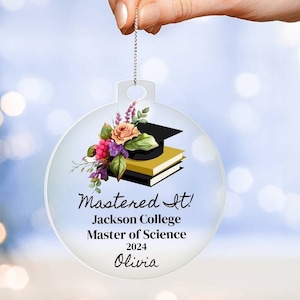 Puede incluir: Un adorno de vidrio transparente con un diseño floral y un gorro de graduación encima de libros. El texto "Mastered It! Jackson College Master of Science 2024 Olivia" está impreso en el adorno.