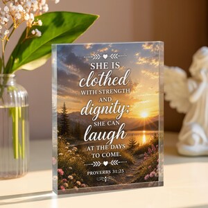 May include: A rectangular clear acrylic plaque featuring a sunset landscape over a lake with mountains and wildflowers. The text reads: SHE IS clothed WITH STRENGTH AND dignity; SHE CAN laugh AT THE DAYS TO COME. PROVERBS 31:25 with heart icons.
