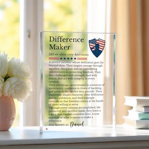 May include: A clear rectangular acrylic plaque featuring the printed text Difference Maker, a phonetic pronunciation, a five-star graphic, and a shield icon with a red, white, and blue American flag pattern. The text describes a service member.