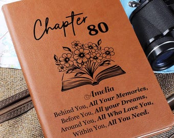 May include: A tan leather-look journal featuring the black text "Chapter 80" above a graphic of an open book with flowers emerging from the pages. Below the graphic, the name "Amelia" is printed, followed by the quote "Behind You, All Your Memories, Before You, All your Dreams, Around You, All Who Love You, Within You, All You Need."