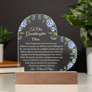 May include: A heart-shaped acrylic plaque with a floral design and a message for a granddaughter. The message reads "To Our Granddaughter Olivia Never forget how much we love you. Wherever you go, you will face many challenges in life. Just stay strong, be confident and do your best. Believe deep in your heart that you are smart and capable of achieving anything your heart desires. When life knocks you down, always choose to get back up. Whenever you feel overwhelmed, know that we are here for you always. We love you always & forever! Love, Mimi & Papa"