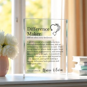 May include: A clear acrylic block featuring the words "Difference Maker" and a definition, accompanied by a floral stethoscope design. The text also includes "Nurse Olivia". The block is on a white surface near a window.