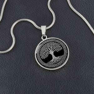 Puede incluir: Collar plateado con un colgante circular. El colgante presenta un diseño de árbol blanco sobre fondo negro, enmarcado por un borde de nudos celtas. La cadena es de estilo serpiente.