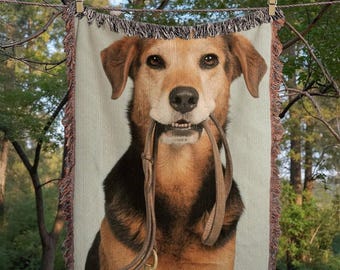 May include: A woven throw blanket featuring a dog holding a leash in its mouth. The dog has brown and black fur and is centered on the cream-colored blanket. The blanket is hanging from a clothesline with wooden clothespins.