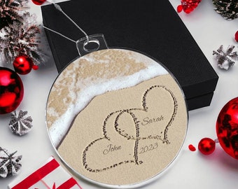 May include: A round, clear ornament with a beach scene and two hearts drawn in the sand. The hearts are labeled "John" and "Sarah" and the year "2023" is written below.