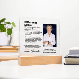Peut inclure: Une plaque acrylique blanche avec une photo en noir et blanc d'une personne. La plaque contient une définition du mot "Difference Maker" et le texte "May you be proud of the work you do and the difference you make!" La plaque porte également le nom "Amanda".