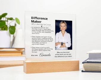 Könnte beinhalten: Eine weiße Acryl-Plakette mit einem Schwarzweißfoto einer Person. Die Plakette enthält eine Definition des Wortes "Difference Maker" und den Text "May you be proud of the work you do and the difference you make!" Die Plakette trägt auch den Namen "Amanda".