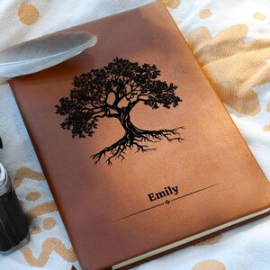 May include: A brown leather journal with a black tree design and the name "Emily" printed on the cover. The journal is closed and resting on a patterned fabric, with a feather nearby.