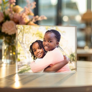 May include: A clear acrylic photo block displaying a color photograph of two young girls embracing. The girls are smiling, and the background is blurred with natural light. The photo block is rectangular and stands upright on a wooden surface.