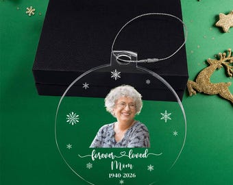 May include: A clear, circular acrylic Christmas ornament featuring a printed photograph of a smiling person with gray hair. The ornament includes white text that reads "forever loved Mom" and "1940-2026," surrounded by small white snowflake graphics.