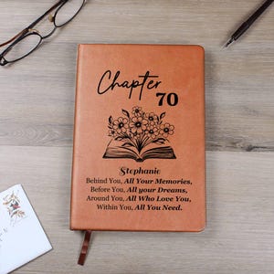 May include: A brown leather journal with the words "Chapter 70" and the name "Stephanie" printed on the cover. The cover also features a floral design above an open book and a quote. A bookmark is attached.