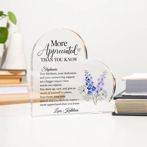 May include: A heart-shaped clear acrylic plaque featuring the printed text "More Appreciated THAN YOU KNOW" followed by a personalised message to Stephanie, a small heart icon, and a floral illustration of blue and purple delphinium flowers.