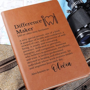 May include: A tan leather journal featuring the text "Difference Maker" and a dental graphic. The journal includes a definition and the name "Olivia". A camera is visible in the background, along with a map.