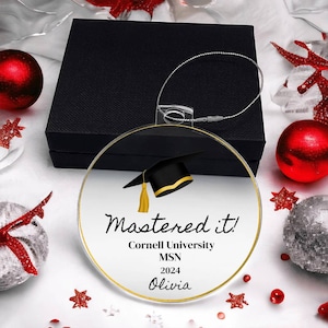 Peut inclure: Une boule de Noël ronde en verre transparent avec un bord doré. La boule présente un bonnet de graduation noir avec un gland doré, et le texte "Mastered it! Cornell University MSN 2024 Olivia".