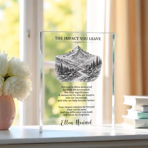 May include: A rectangular clear acrylic plaque featuring a black ink illustration of a mountain range and forest. The text reads: THE IMPACT YOU LEAVE, followed by an inspirational quote and the name Ellen Howard printed in black lettering.