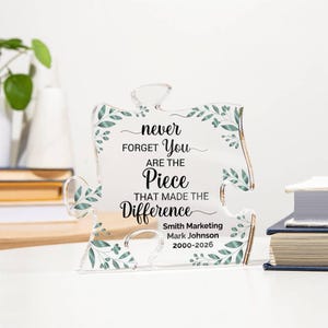 Könnte beinhalten: Klarer Acryl-Puzzleteil mit dem Text "never forget you are the piece that made the difference". Das Teil hat grüne Blätterakzente und die Namen "Smith Marketing" und "Mark Johnson 2000-2026".