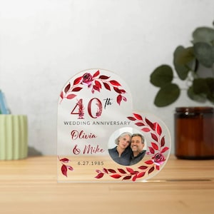 May include: A clear acrylic heart-shaped anniversary photo frame. The frame features a photo of a couple, with the text "40th Wedding Anniversary" and the names "Olivia & Mike" with the date "6.27.1985". The frame is decorated with red floral accents.