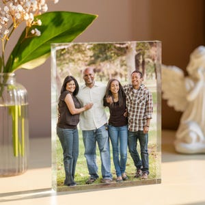 May include: A clear acrylic photo block featuring a family portrait. The image shows a family of four smiling outdoors. The block is rectangular and stands upright on a light-coloured surface, with a vase of flowers to the left.