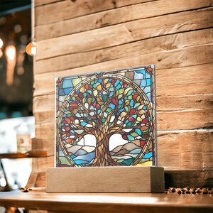 Könnte beinhalten: Ein Buntglasfenster mit einem Baumdesign mit bunten Blättern, das in einem kreisförmigen Design angeordnet ist. Das Kunstwerk ist auf einem rechteckigen Holzsockel montiert. Das Panel verwendet Blau-, Grün-, Gelb- und Rottöne und schafft ein lebendiges Dekorationsstück.