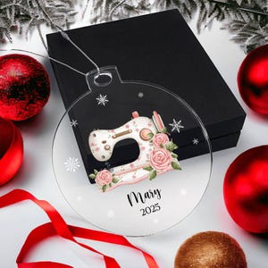 May include: Clear, round Christmas ornament featuring a sewing machine design with pink roses and snowflakes. The ornament has the name "Mary" and the year "2025" printed on it. The ornament is displayed with red and gold Christmas ornaments and a black gift box.