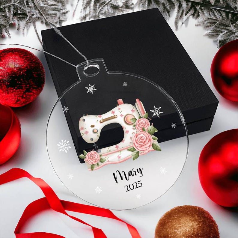 May include: Clear, round Christmas ornament featuring a sewing machine design with pink roses and snowflakes. The ornament has the name "Mary" and the year "2025" printed on it. The ornament is displayed with red and gold Christmas ornaments and a black gift box.