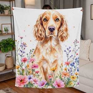 Può includere: Una morbida coperta in pile bianco con un'illustrazione ad acquerello di un cocker spaniel dorato seduto in un campo di fiori selvatici colorati.