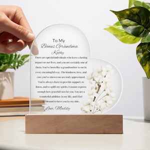 May include: A heart-shaped glass plaque with a floral design and a message to a bonus grandma. The plaque reads "To My Bonus Grandma Kathy There are special individuals who leave a lasting impact on our lives, and you are certainly one of them. You've been like a grandmother to me in every meaningful way. The kindness, love, and care you've shown me are truly appreciated. You're always there to provide support, to listen, and to uplift my spirits. I cannot express enough how grateful I am for you. You are a wonderful addition in my life, and I feel so blessed to have you by my side. Love, Maddy"