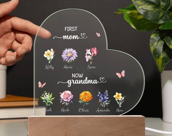 May include: A heart-shaped acrylic sign with the text "First mom" and "Now grandma" with a floral design and names of children. The sign is lit up with a warm white light and sits on a wooden base.