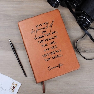 May include: A tan leather journal with the inscription "MAY YOU be proud of THE WORK YOU DO, THE PERSON YOU ARE, AND THE DIFFERENCE YOU MAKE." and the name "Samantha". A black pen and binoculars are also visible.