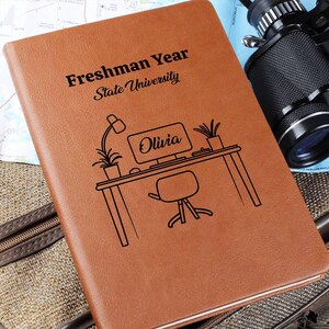 May include: A brown leather journal with the text "Freshman Year State University" and a desk illustration with the name "Olivia". The journal is on a map and a suitcase.
