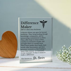 May include: A clear acrylic block with the words "Difference Maker" and a definition, along with a medical symbol. The text describes a doctor's care and commitment. The block is on a white surface with a wooden heart and flowers.