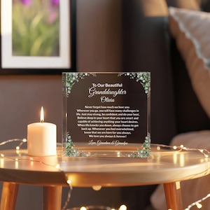 May include: A clear acrylic block with a heartfelt message for a granddaughter, adorned with floral accents. A lit white candle and string lights are on a wooden table, creating a warm, sentimental scene.