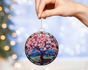May include: A circular hanging ornament featuring a stained glass style design of a blooming cherry blossom tree. The tree has a dark brown trunk and branches with numerous pink flowers, set against a blue sky and green landscape background.