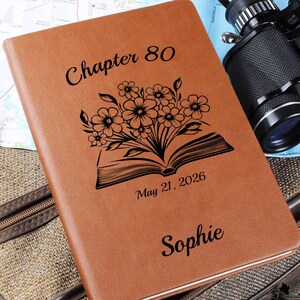 May include: A tan notebook with a black engraved design featuring an open book with flowers emerging from the pages. The text reads Chapter 80, May 21, 2026, and the name Sophie. The cover has a smooth, leather-like texture and rounded corners.