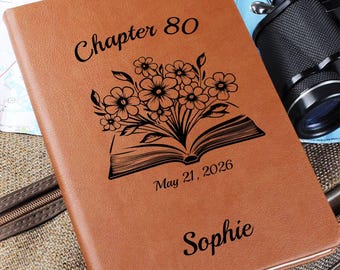 May include: A tan notebook with a black engraved design featuring an open book with flowers emerging from the pages. The text reads Chapter 80, May 21, 2026, and the name Sophie. The cover has a smooth, leather-like texture and rounded corners.