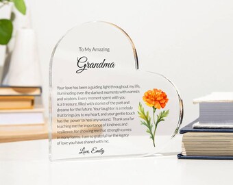 May include: A heart-shaped clear acrylic plaque featuring the text To My Amazing Grandma, a heartfelt message about love and wisdom, and a printed orange marigold flower. The plaque is signed with the name Emily at the bottom in black script.