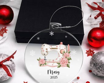 May include: A clear, round Christmas ornament featuring a sewing machine design with pink roses and the name "Mary" and the year "2025". The ornament has a silver hanging wire and is surrounded by holiday decorations.