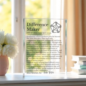 Könnte beinhalten: Ein klarer Acrylblock mit den Worten "Difference Maker" und einer Definition. Der Block zeigt eine Grafik von Händen, die ein Herz halten. Der Text beschreibt die Freundlichkeit und den Einfluss eines Freiwilligen. Enthält auch den Namen "Kate Foster".